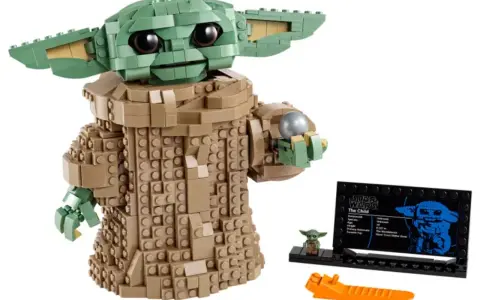 Figurka Grogu z klocków LEGO, znana z gier LEGO Star Wars, trzyma srebrną kulę. Obok stoi mała figurka i tabliczka informacyjna.