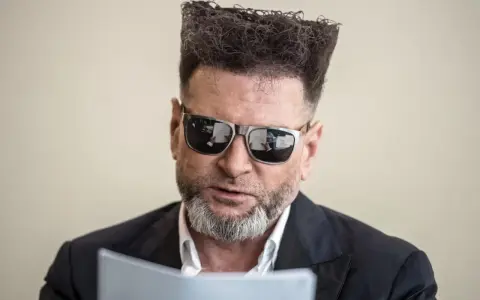 Detektyw Rutkowski is back! Z charakterystyczną fryzurą i okularami, czyta dokumenty.