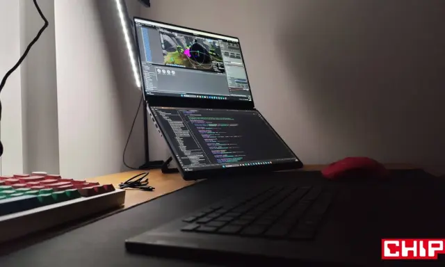 Na biurku laptop z dwoma ekranami, na górnym widać scenę w Unreal Engine 6, na dolnym kod. Obok klawiatura i czerwona myszka.